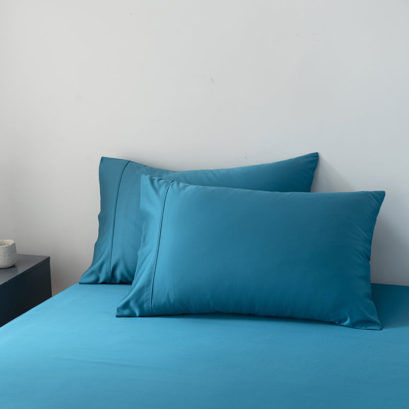 Teal bamboo bed sheets and pillows on a bed against a plain wall.